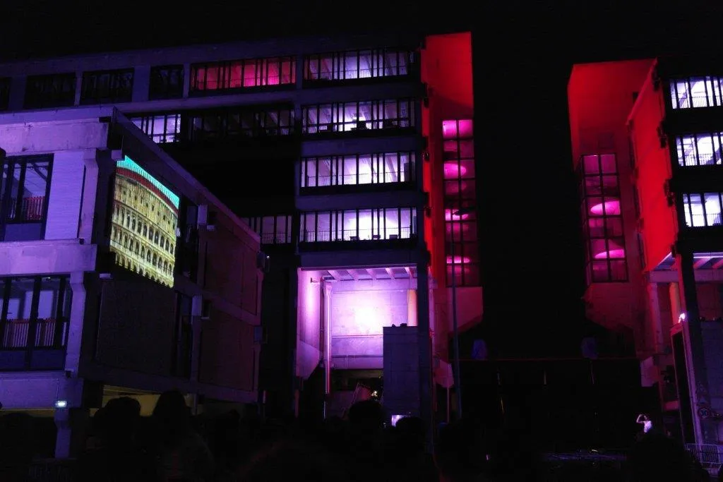 A courtyard bathed in red and violet light with a projected monument