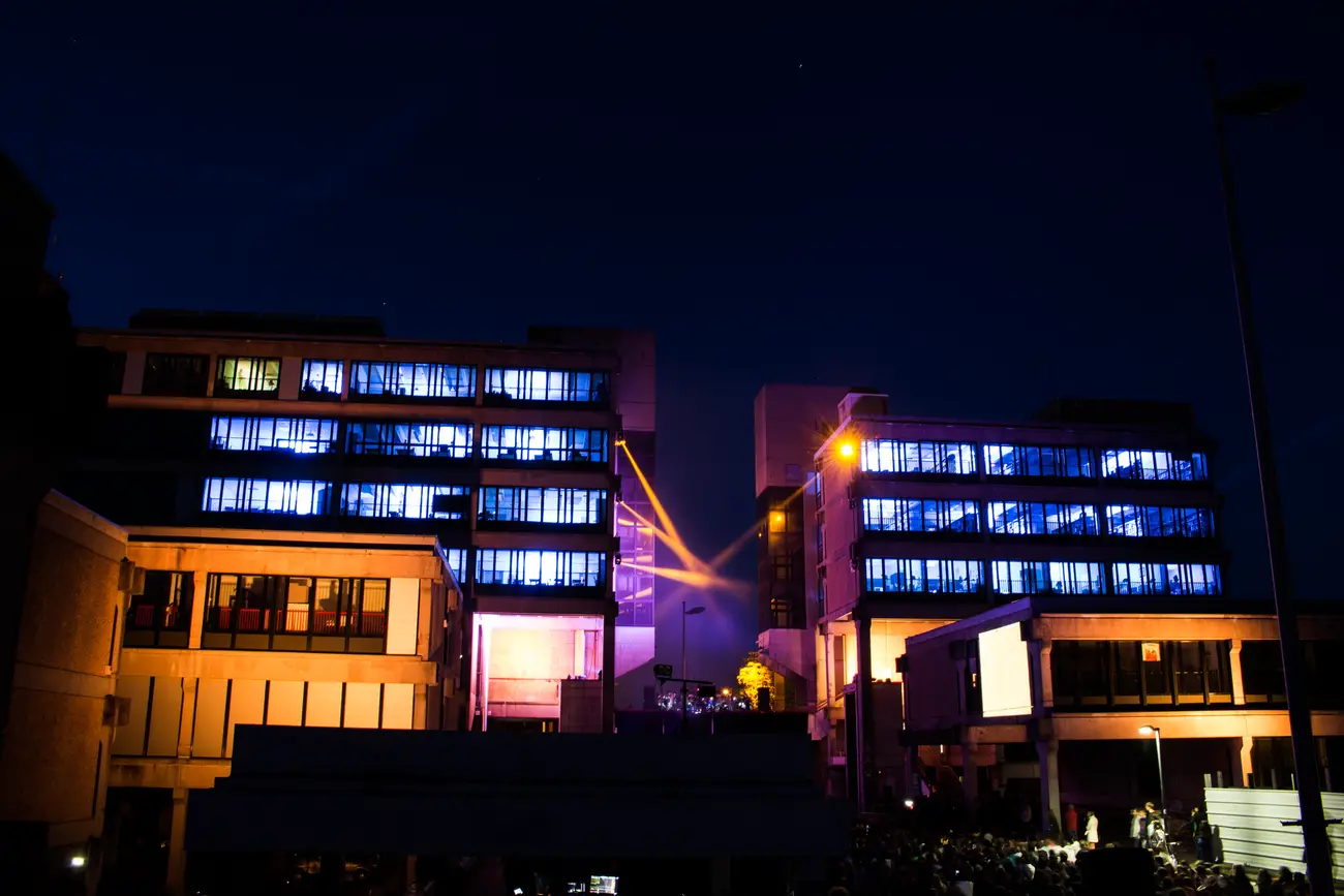 Two campus buildings glowing blue and amber at night, a spotlight beam crossing between them